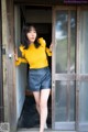 A woman standing in front of a door wearing a yellow sweater and black shorts.